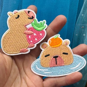 May include: Two embroidered capybara patches. One holds a strawberry, the other floats in blue water with an orange on its head. The patches have a white border and are held in a hand. The capybaras are cartoon-style.