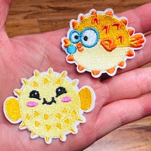 May include: Two embroidered patches featuring cartoon pufferfish. One has blue eyes and orange accents, the other a smiling face with pink cheeks. Both are yellow with white and orange trim.