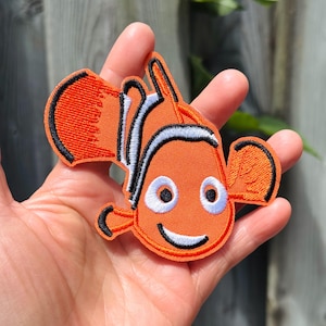 May include: An embroidered patch shaped like a clownfish with bright orange thread, white stripes outlined in black, and two large white eyes with black pupils. The fish has a curved white mouth and black-edged fins, creating a detailed design.