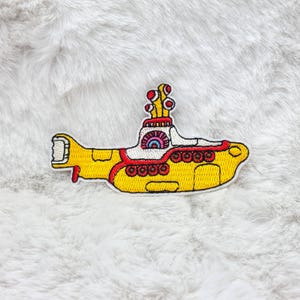 May include: Embroidered patch depicting a yellow submarine with red and white detailing. The patch has a cartoon-like design, with detailed stitching and a playful aesthetic. The submarine has a unique, stylized design, perfect for adding to clothing or accessories.