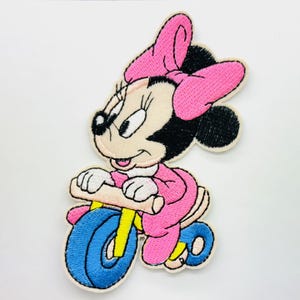 Może przedstawiać: Naszywka haftowana do prasowania z postacią kreskówkową Minnie Mouse jadącej na różowym trójkołowym rowerze z niebieskimi kołami. Minnie ma na sobie różową sukienkę i różową kokardkę.