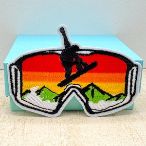 Op de afbeelding: Geborduurde patch met een snowboarder in de lucht in een skibril. De lenzen van de bril tonen een zonsondergang met oranje, gele en rode tinten boven groene bergtoppen. De patch heeft een witte rand.