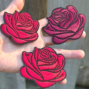 May include: Three embroidered rose patches featuring vibrant magenta and deep red thread with black stitched outlines. Each patch displays a detailed floral design with layered petals, creating a textured appearance suitable for fabric application.