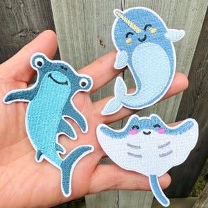 Può includere: Tre toppe ricamate raffiguranti creature marine: uno squalo martello, un narvalo e una manta. Le toppe sono principalmente blu e bianche con dettagli neri e gialli. Il narvalo ha un corno e la manta ha le guance rosa.