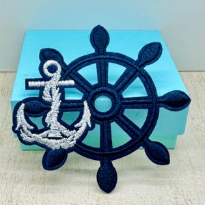 May include: A navy blue embroidered patch with a silver anchor and a ship's wheel. The anchor is in the center of the patch and the wheel is around it.