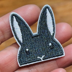 May include: Embroidered patch depicting a rabbit's head. The design uses dark blue and iridescent thread with white detailing in the ears and a white border. A charming, whimsical patch.