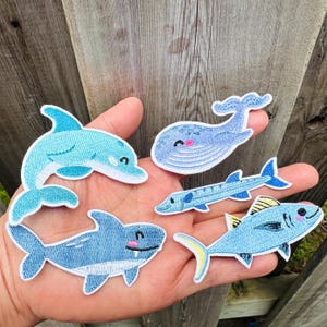 Peut inclure: Plusieurs écussons brodés représentant des créatures marines. Les écussons incluent un dauphin, une baleine, un requin, un barracuda et un thon. Chaque écusson est bleu clair avec des détails blancs et jaunes et un visage souriant.