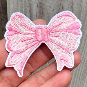 May include: Embroidered pink bow patch with a white border. The bow features detailed stitching and a classic bow shape. Suitable for clothing, accessories, or craft projects.