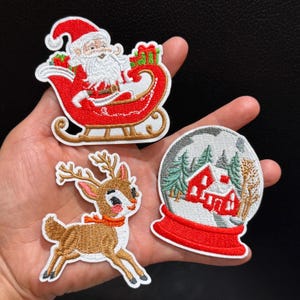 Könnte beinhalten: Drei bestickte Aufnäher mit Weihnachtsmotiven: ein rot-weißer Weihnachtsmann im Schlitten mit grünen Geschenken, ein braunes Rentier mit rotem Schal und eine Schneekugel mit einem roten Haus, umgeben von grünen Tannenbäumen.