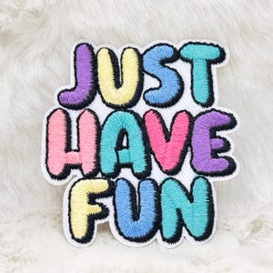 May include: Embroidered patch featuring the phrase "JUST HAVE FUN" in bubble letters. The letters are in various colours, including purple, yellow, pink, teal, and blue, with black outlines. The patch is set against a white backdrop.