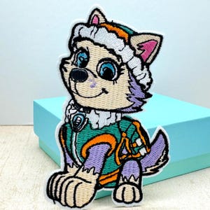 May include: Embroidered patch of a cartoon dog wearing a purple and green winter outfit with a white hat. The dog has blue eyes and a pink nose.