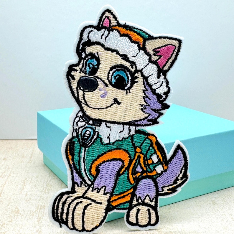May include: Embroidered patch of a cartoon dog wearing a purple and green winter outfit with a white hat. The dog has blue eyes and a pink nose.