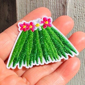 Op de afbeelding: Geborduurde patch met een groen grasrokontwerp met een witte geschulpte rand. De patch is versierd met roze en gele bloemaccenten, waardoor een tropisch thema ontstaat. De patch is waarschijnlijk voor kleding of accessoires.