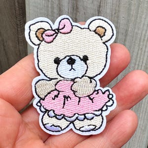 May include: An embroidered patch featuring a cream-colored teddy bear with a light blue muzzle and black stitched facial features. The bear wears a pink dress with a ruffled hem and a matching pink bow on its head, outlined in white thread.