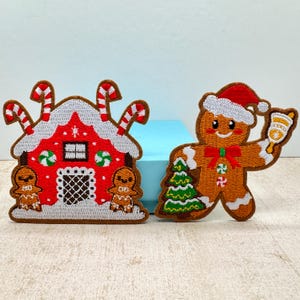 Op de afbeelding: Twee kerst-thema geborduurde patches. Eén is een rood peperkoekhuisje met witte glazuur en zuurstok accenten. De andere is een peperkoekmannetje met een kerstmuts, met een kerstboom en een bel.