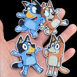 Op de afbeelding: Vier geborduurde patches met stripfiguurhonden. Twee patches tonen een blauwe hond met zwarte oren en een gele snuit, terwijl twee patches een roodbruine hond met een crèmekleurige buik en gezicht afbeelden, allemaal met grote ogen.