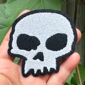 May include: A white skull patch with black eye sockets, nose, and outline. The patch has a textured appearance and is likely made of fabric. The design is a simple, yet bold, representation of a skull.