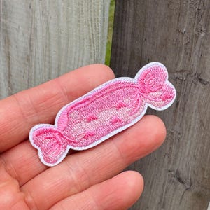 Può includere: Una toppa di caramella rosa ricamata con un bordo bianco. La caramella è avvolta in un classico design a torsione. La toppa è tenuta in mano da una persona, con una staccionata di legno sullo sfondo. Perfetta per abbigliamento o accessori.