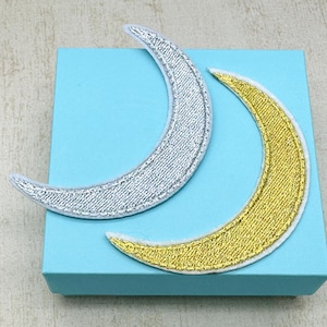 May include: Two crescent moon shaped iron-on patches, one silver and one gold, on a light blue background.