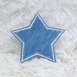 Può includere: Toppa a forma di stella in denim blu con cuciture bianche sui bordi. La toppa è posta su uno sfondo morbido, bianco e strutturato. La stella ha un classico design a cinque punte.