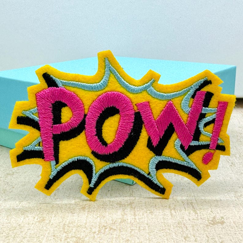 Op de afbeelding: Een gele viltpatch met een zwarte omtrek en een roze "POW!" in het midden. De patch is gevormd als een comicboek-explosie.