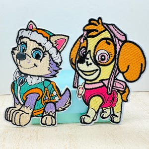 May include: Two embroidered patches featuring characters from the animated series PAW Patrol. The patch on the left features a purple and white husky wearing a green and orange jacket. The patch on the right features a yellow and orange dog wearing a pink and white jacket.