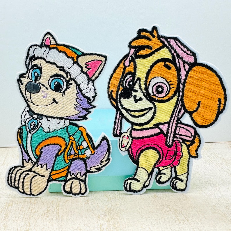 May include: Two embroidered patches featuring characters from the animated series PAW Patrol. The patch on the left features a purple and white husky wearing a green and orange jacket. The patch on the right features a yellow and orange dog wearing a pink and white jacket.