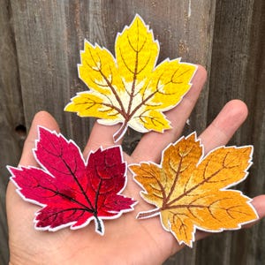 Pode incluir: Três patches bordados de folhas de bordo em cores de outono: amarelo, vermelho e laranja. Cada folha tem costuras detalhadas, uma borda branca e um caule marrom. As folhas são exibidas em um fundo de madeira.