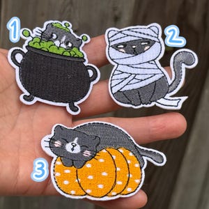 Könnte beinhalten: Drei bestickte Aufnäher mit Katzenmotiven. Nummer 1 zeigt eine schwarz-weiße Katze in einem schwarzen Kessel mit grünen Blasen. Nummer 2 ist eine schwarze Katze, die in weiße Bandagen gewickelt ist. Nummer 3 ist eine schwarz-weiße Katze auf einem orangefarbenen Kürbis.