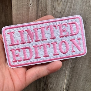 May include: A rectangular patch embroidered with the words "LIMITED EDITION" in pink on a white background. The patch has a pink border. The patch is held against a wooden surface.