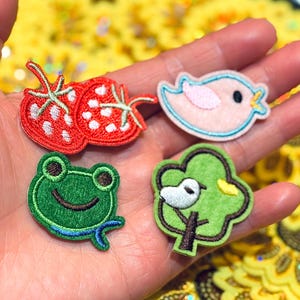 Op de afbeelding: Vier geborduurde patches: twee rode aardbeien, een roze vogel, een groene kikker en een groene boom met een witte vogel. De patches hebben kleurrijke stiksels en zijn waarschijnlijk voor kleding of knutselen.