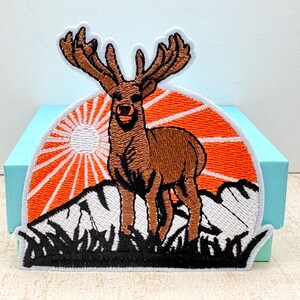 May include: Embroidered patch depicting a brown deer with large antlers, set against a mountain range and an orange sunset. The patch has a white border. The background is light blue.