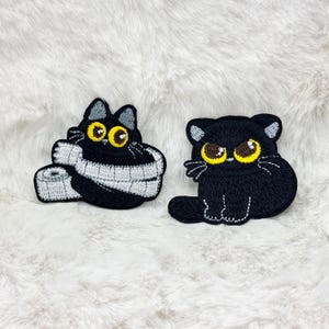 May include: Two embroidered patches of black cats. One cat is wrapped in a grey and white measuring tape, and the other is sitting. Both cats have yellow eyes and grey accents.