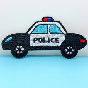 Puede incluir: Parche bordado de un coche de policía. El coche es negro con un panel de puerta blanco que dice "POLICÍA" en letras negras en negrita. Las ventanas son de color azul claro. El coche tiene una luz roja y azul en la parte superior y luces rojas y azules en la parte delantera.