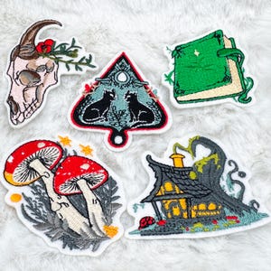 May include: A selection of embroidered patches with diverse designs. Includes a skull with horns and flowers, two black cats in a triangle, a green book, red mushroom, and a spooky house. Each patch has a white border.
