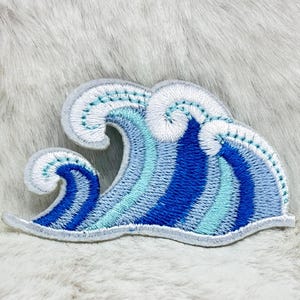 May include: An embroidered patch shaped like a stylised ocean wave. The design features layered shades of dark blue, medium blue, and light blue thread, with white stitching outlining the crests of the waves and the outer edges of the patch.
