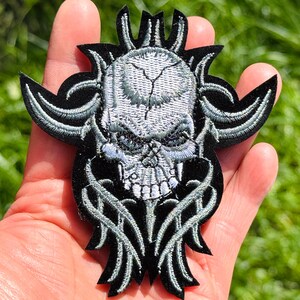 May include: A black embroidered patch featuring a silver-grey skull design with intricate, pointed, and curved tribal-style patterns extending from the sides and bottom. The embroidery is detailed with light grey stitching on a solid black fabric base.