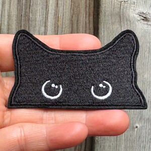May include: Black embroidered cat patch featuring white crescent-shaped eyes. The patch has a rectangular shape with rounded corners and a black border. This patch is ideal for adding to clothing, bags, or other fabric items.