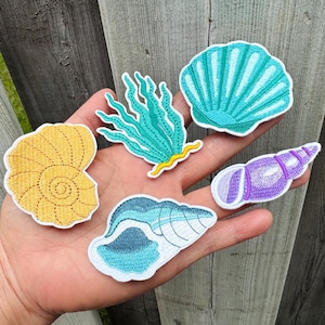 以下が含まれることがあります： さまざまな海洋生物のデザインが施された刺繍パッチのコレクション。パッチには、黄色の渦巻き貝、ターコイズの海藻、ターコイズのホタテ貝、青と白の貝、紫色の巻貝が含まれています。各パッチには白い縁取りがあります。