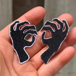 Op de afbeelding: Twee geborduurde patches in de vorm van handen die een oké-gebaar maken. Elke patch is gemaakt van zwart garen dat het silhouet van de hand vormt, met een opvallende witte geborduurde rand die de vingers en handpalm omlijnt voor contrast.