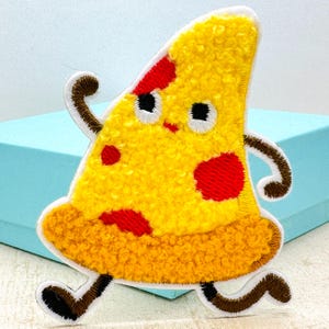 Puede incluir: Parche bordado con una porción de pizza de dibujos animados con una cara sonriente, pepperoni rojo y brazos y piernas marrones. La porción de pizza es amarilla con un aspecto texturizado y un borde blanco. El parche está diseñado para que parezca que la porción de pizza está corriendo.