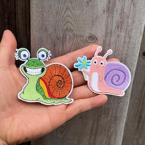 Puede incluir: Dos parches bordados con caracoles de dibujos animados. Uno es verde con una concha naranja y una gran sonrisa. El otro es rosa con una concha morada y sostiene una flor azul. Fondo de madera.