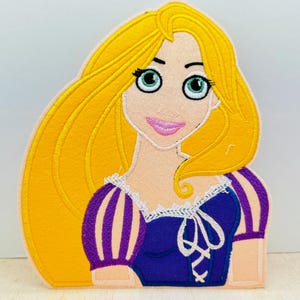 Op de afbeelding: Gestikt vilt patch van een cartoon prinses met lang blond haar, die een paars-witte jurk draagt.