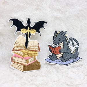 Könnte beinhalten: Gestickte Aufnäher mit Drachen und Büchern. Ein Aufnäher zeigt einen schwarzen Drachen mit goldenen Akzenten über einem Stapel Bücher. Ein anderer zeigt einen grauen Drachen, der ein Buch auf einem blauen Kissen liest.