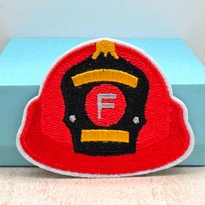 May include: Embroidered patch of a red firefighter helmet with a black faceplate and yellow accents. The helmet features a white border and a silver letter "F" in the center.