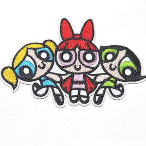 May include: Embroidered patch featuring the Powerpuff Girls. The patch depicts Blossom, Bubbles, and Buttercup in their signature colours: red, blue, and green. The cartoon characters are smiling and holding hands, with a white border.