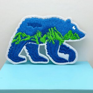 May include: Embroidered patch in the shape of a bear, featuring a blue body with green mountain details. The patch has a white border and is placed on a light blue surface. Ideal for jackets, bags, or other fabric items.
