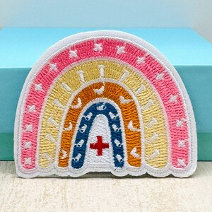 May include: Embroidered rainbow patch with a white border. The rainbow features layers of pink, yellow, orange, and blue, with a red cross symbol at the bottom. Small white star-like shapes are embroidered throughout the design.