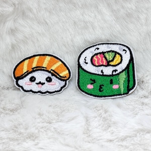 May include: Two embroidered patches featuring sushi designs. One patch depicts a piece of nigiri sushi with orange and white details. The other patch shows a green maki roll with colorful fillings. Both patches have cute, smiling faces.