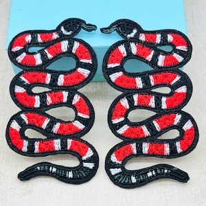 May include: Two embroidered snake patches. Each patch features a red and white striped pattern on a black background. The snake's head is at the top, with a curved body. The patches are likely for clothing or accessories.